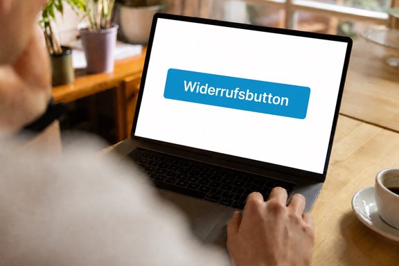 Blick über die Schulter eines Mannes am Schreibtisch. Vor ihm steht ein geöffneter Laptop. Darauf zu sehen ist ein großer blauer Button, auf dem steht "Widerrufsbutton".