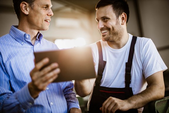 Mann im Hemd zeigt Mann in Arbeitskleidung etwas am Tablet
